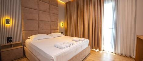 A modern hotel room with a large bed, wooden flooring, and a view of the outdoors through curtains.