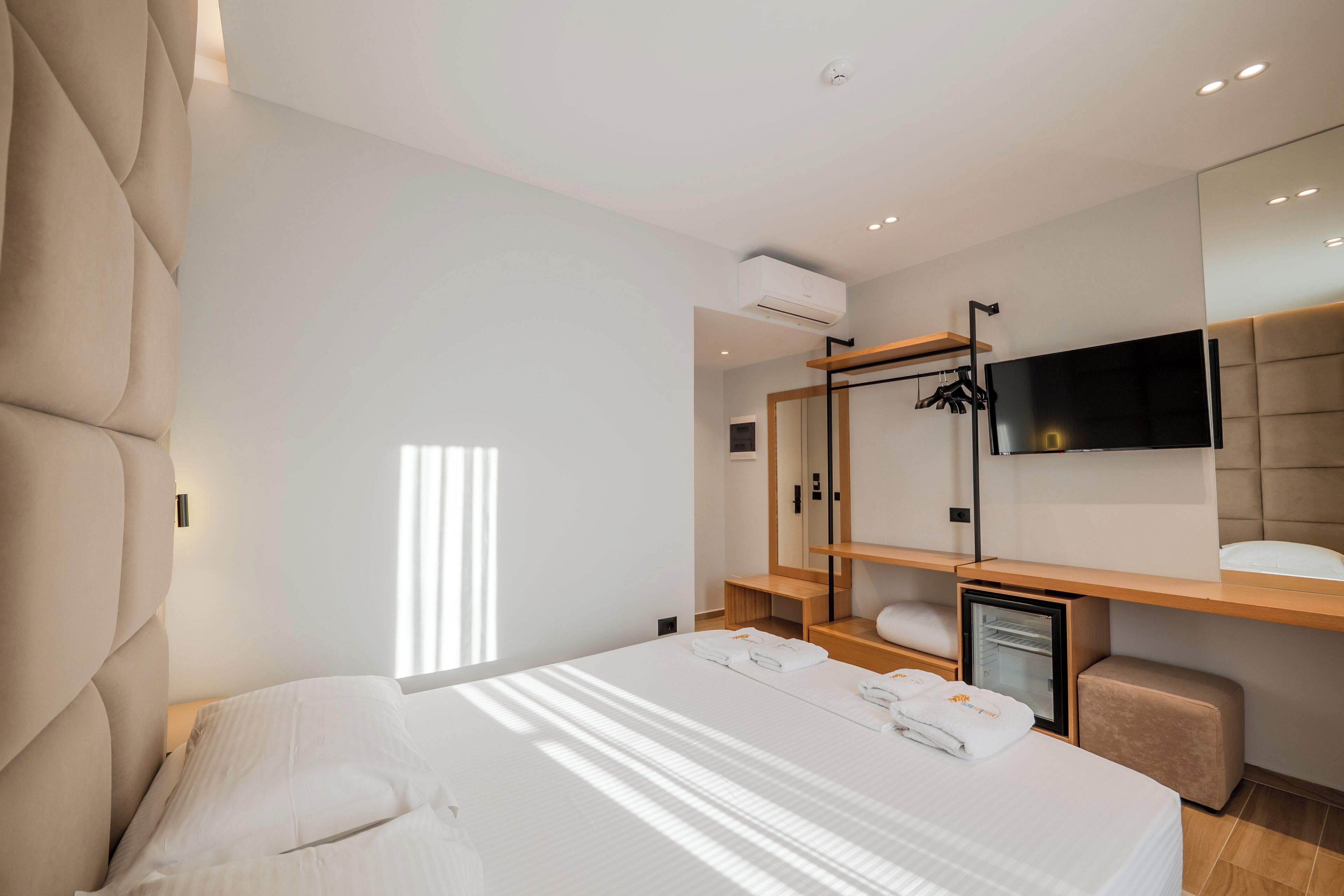 A modern hotel room with a large bed, a TV mounted on the wall, a small seating area, and a built-in air conditioning unit.