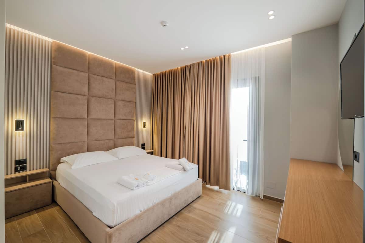 A modern hotel room with a large bed, a wooden nightstand, a flat-screen TV, and a large window with curtains.