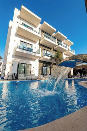 Un edificio moderno con piscina que incluye una fuente, asientos al aire libre y un cielo azul despejado.