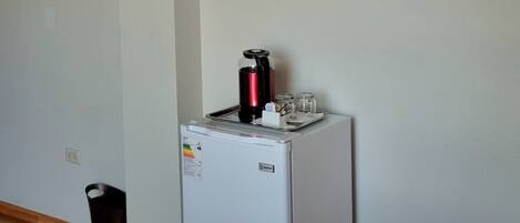 Habitación con televisión de pantalla plana montada en la pared, un refrigerador pequeño, un basurero y un tapete azul en el suelo.