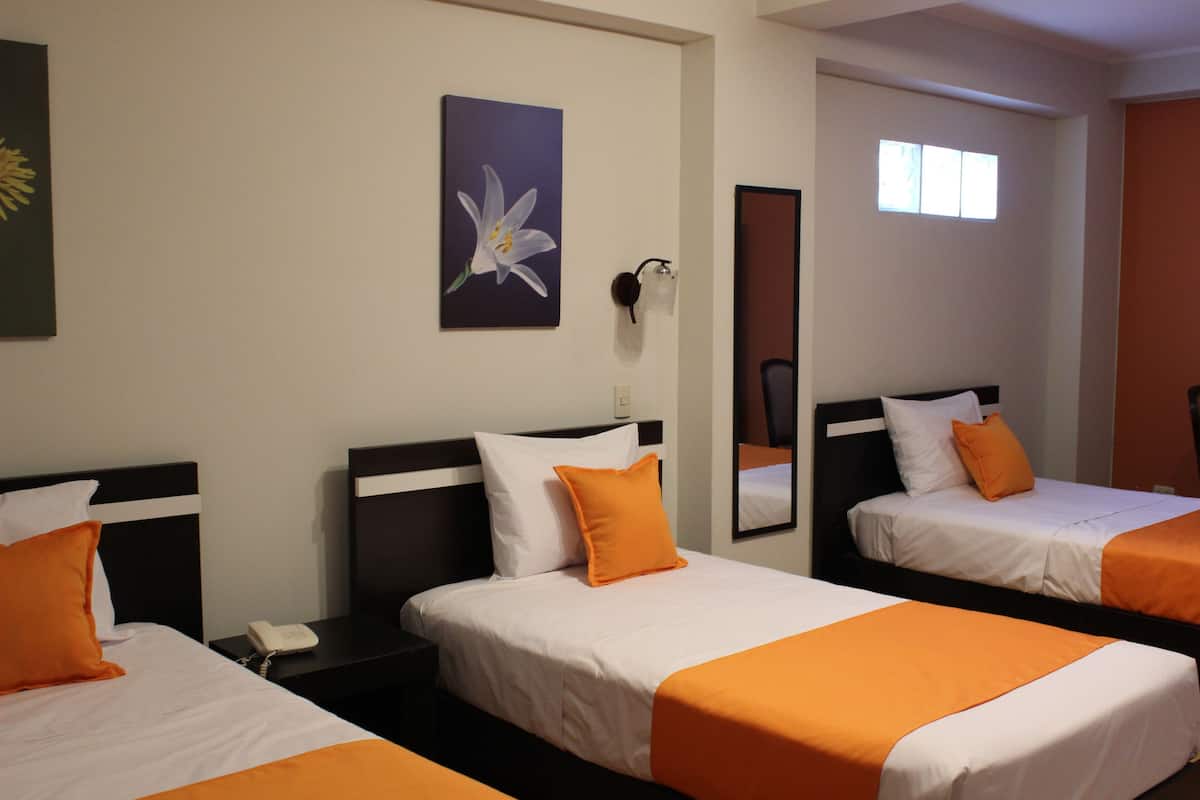 Habitación de hotel con dos camas, cada una con ropa blanca y detalles naranja, una mesita de noche con un teléfono, una lámpara de pared, un cuadro con una pintura floral y una ventana con luz natural.