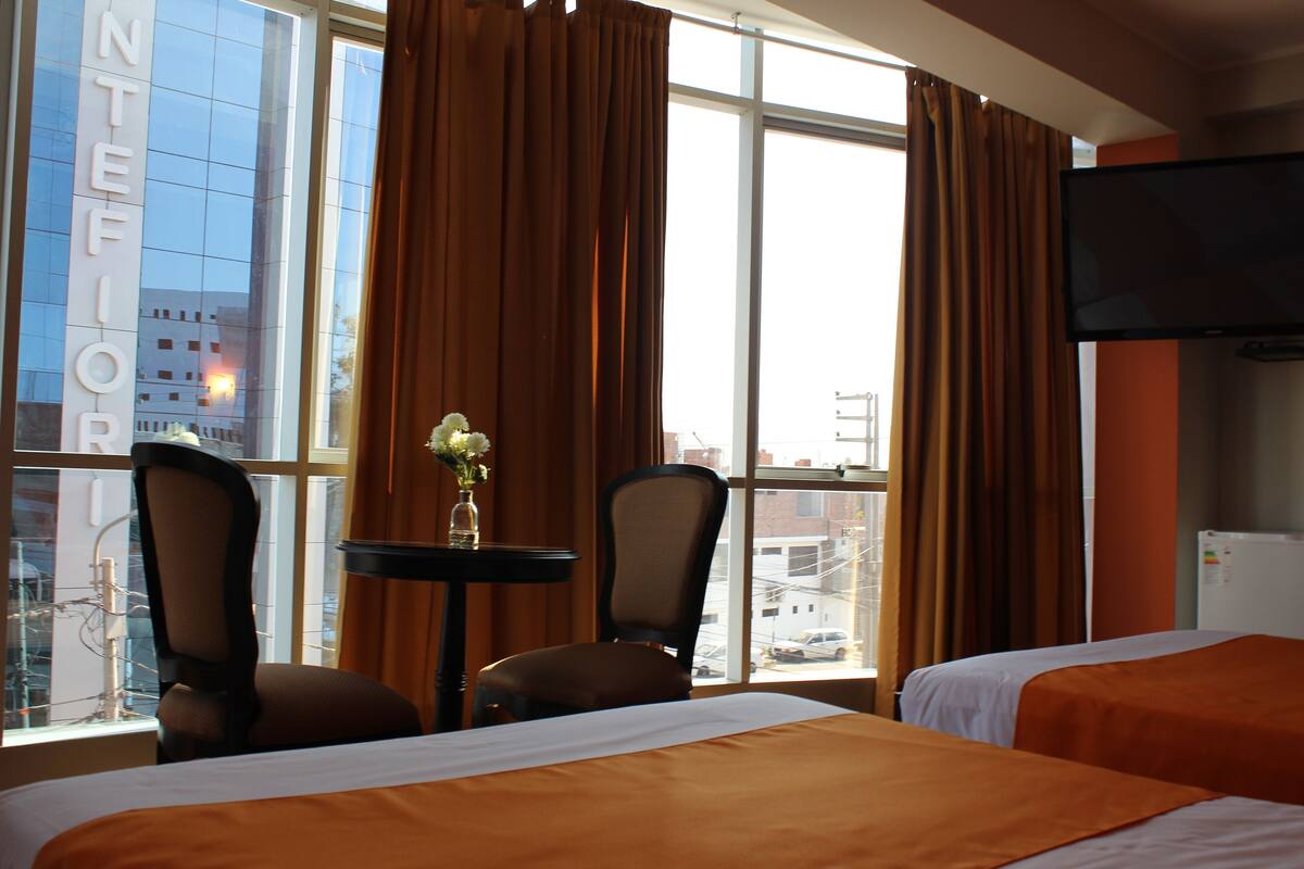 Habitación de hotel con dos camas, una mesita con un florero, dos sillas, un televisor y una ventana con cortinas.
