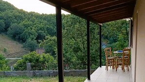 A covered patio with tiled flooring, a table and chairs, and a view of a green landscape with trees and hills. - Billy s House Vlachava-kalambaka (Kalabaka)