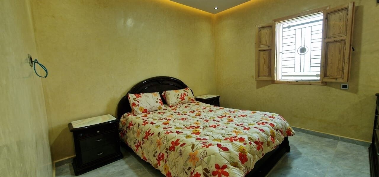 A bedroom with a bed, nightstands, a dresser, a window with wooden shutters, and a wall-mounted light fixture.