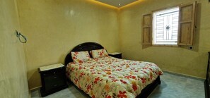 A bedroom with a bed, nightstands, a dresser, a window with wooden shutters, and a wall-mounted light fixture.