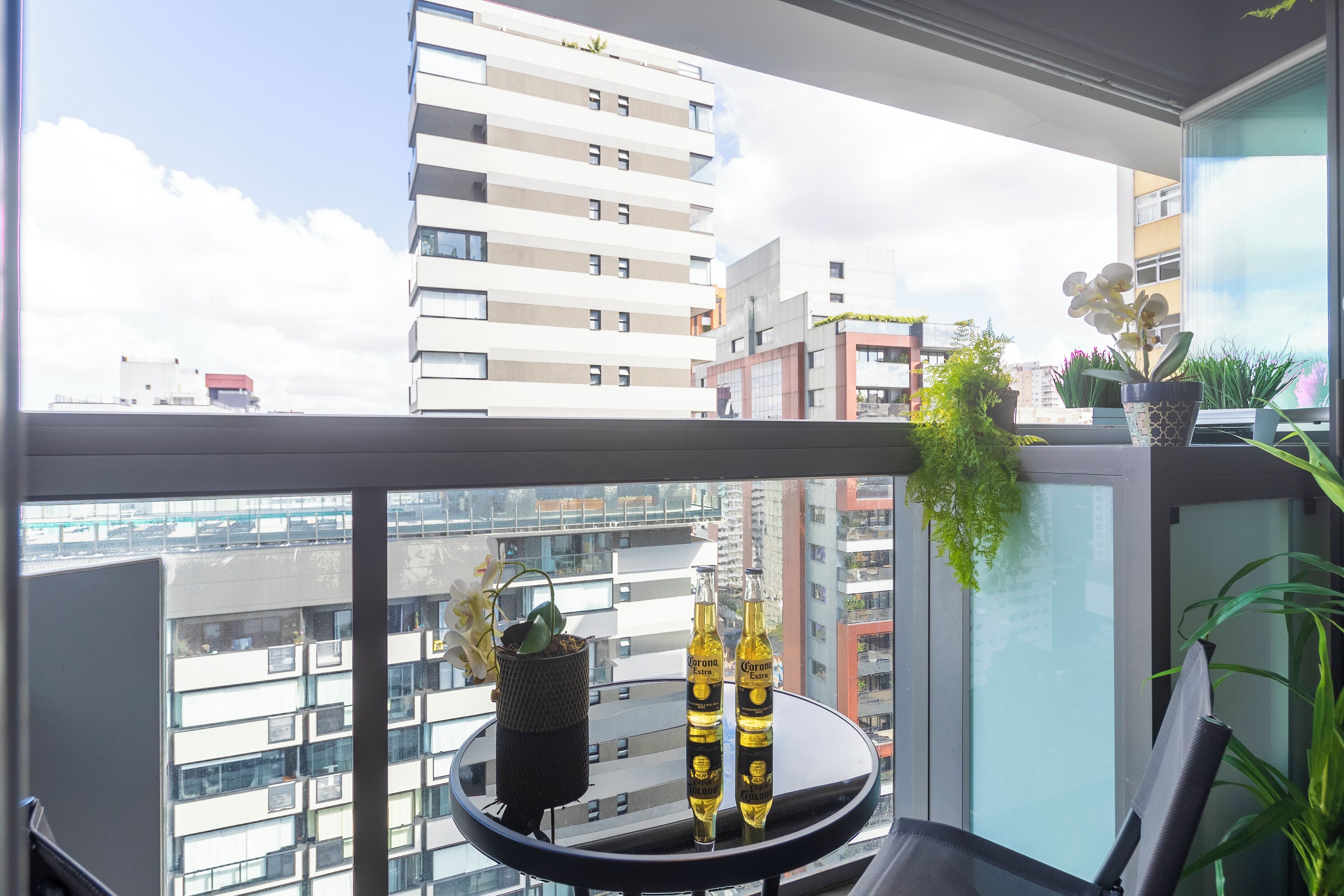 A balcony with a view of a cityscape, a small table with two bottles of Corona beer, and a potted orchid.