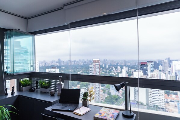 A modern office with a large window, a desk with a laptop, a lamp, and decorative plants.