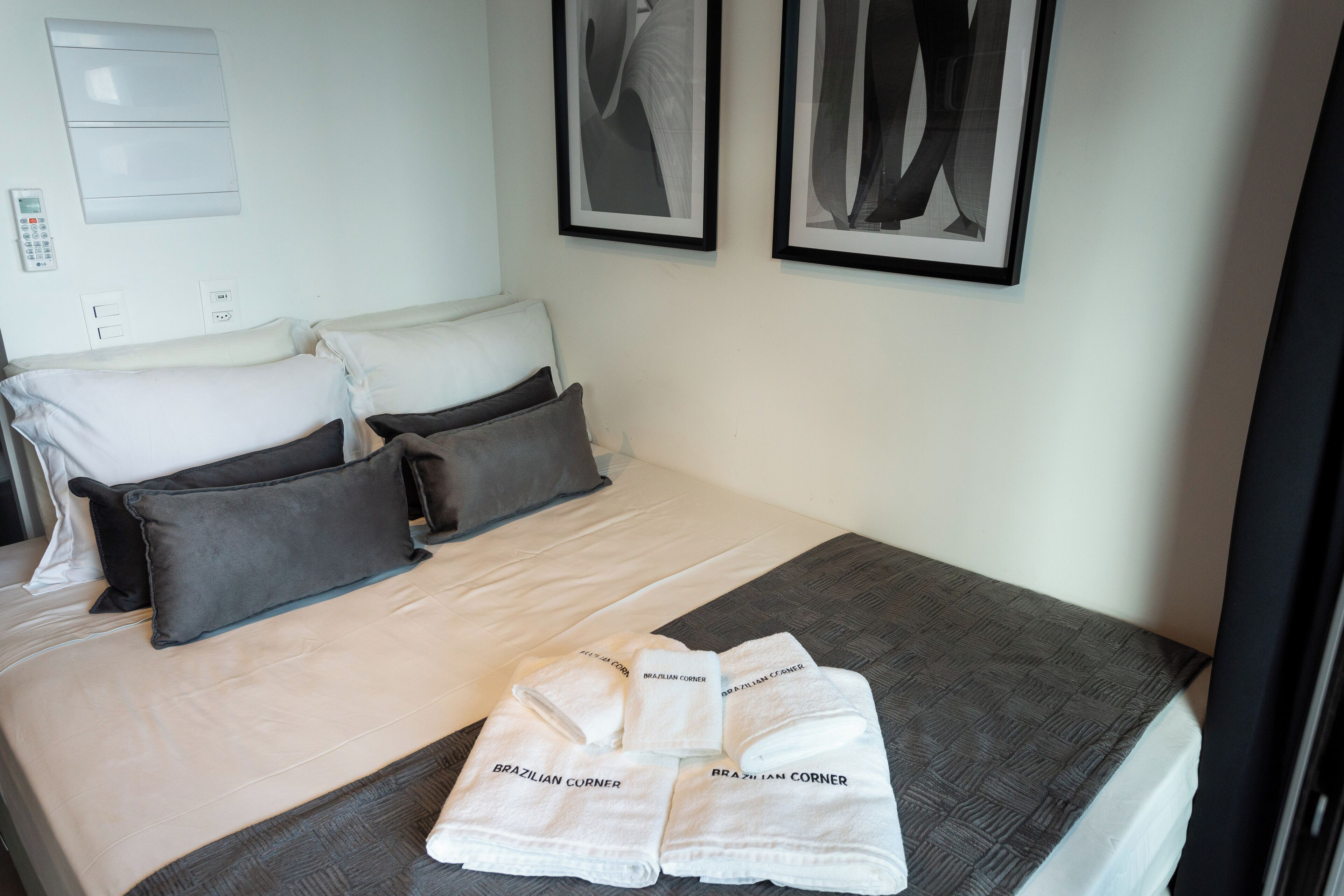 Un lit bien fait, avec une literie blanche et grise, un ensemble de serviettes portant la marque « Brazilian Corner », et deux tableaux encadrés accrochés au mur.