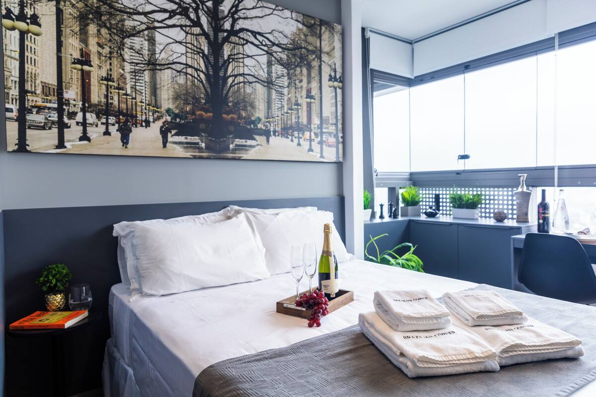 Ein Hotelzimmer mit Bett, einem Fenster mit Blick auf Gebäude, einem Bild an der Wand und einer Champagnerflasche auf dem Bett.