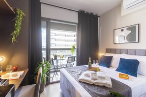 A hotel room with a bed, a dining table with chairs, a balcony with a view of buildings, and a window with curtains.