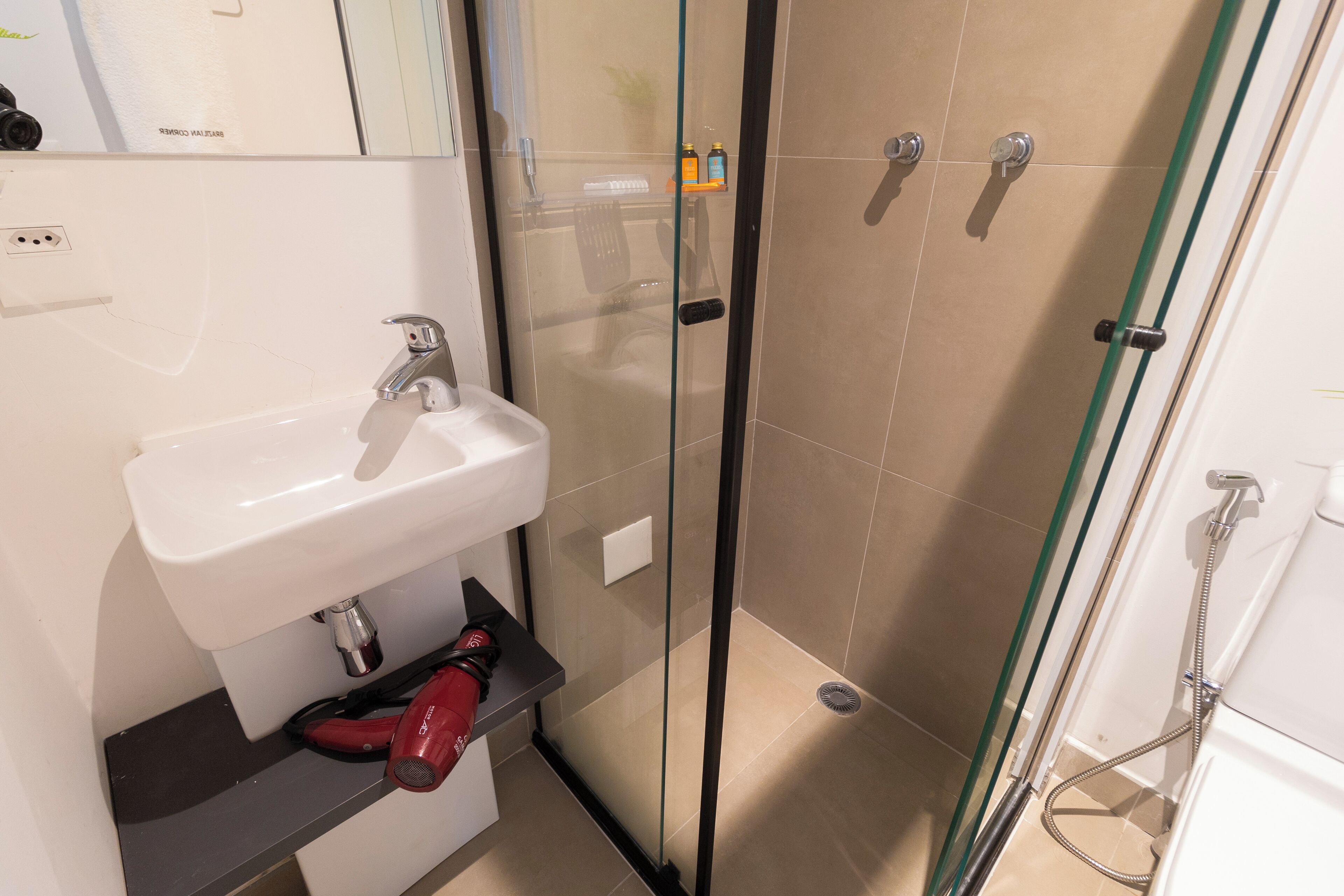 Ein Badezimmer mit einer gläsernen Duschkabine, einem weißen Waschbecken und einem roten Fön auf einem Regal.