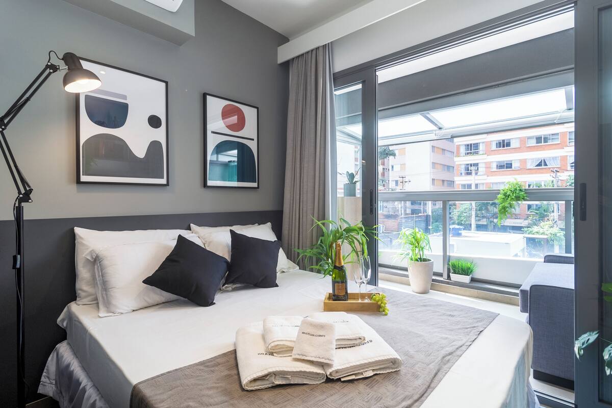Ein modernes Hotelzimmer mit einem großen Bett, einer Nachttischlampe, einem Blumentopf und Blick auf die Stadt durch große Fenster.