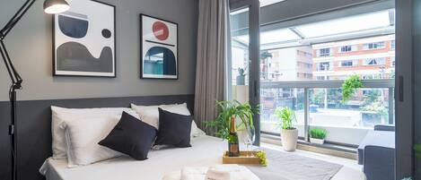 Una moderna camera d'albergo con un letto grande, una lampada da comodino, una pianta in vaso e vista sulla città attraverso ampie finestre.