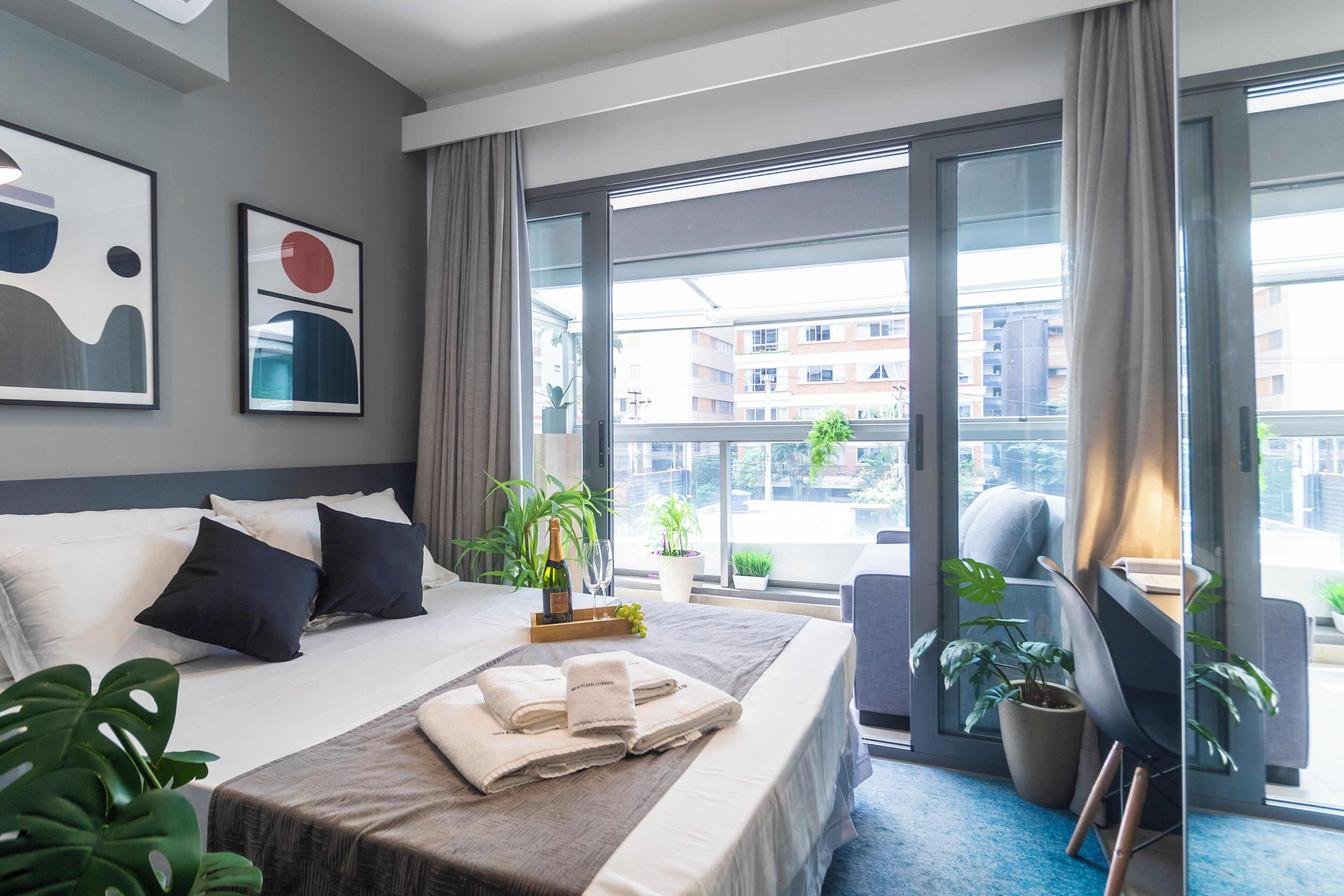 A hotel room with a large bed, a desk, a chair, and a view of the city through large windows.