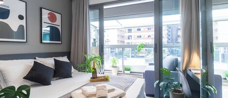 A hotel room with a large bed, a desk, a chair, and a view of the city through large windows.