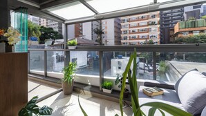 A balcony with a view of surrounding buildings, potted plants, and a sofa with a book. - Oscar Freire - Condomínio Glass VN (São Paulo)