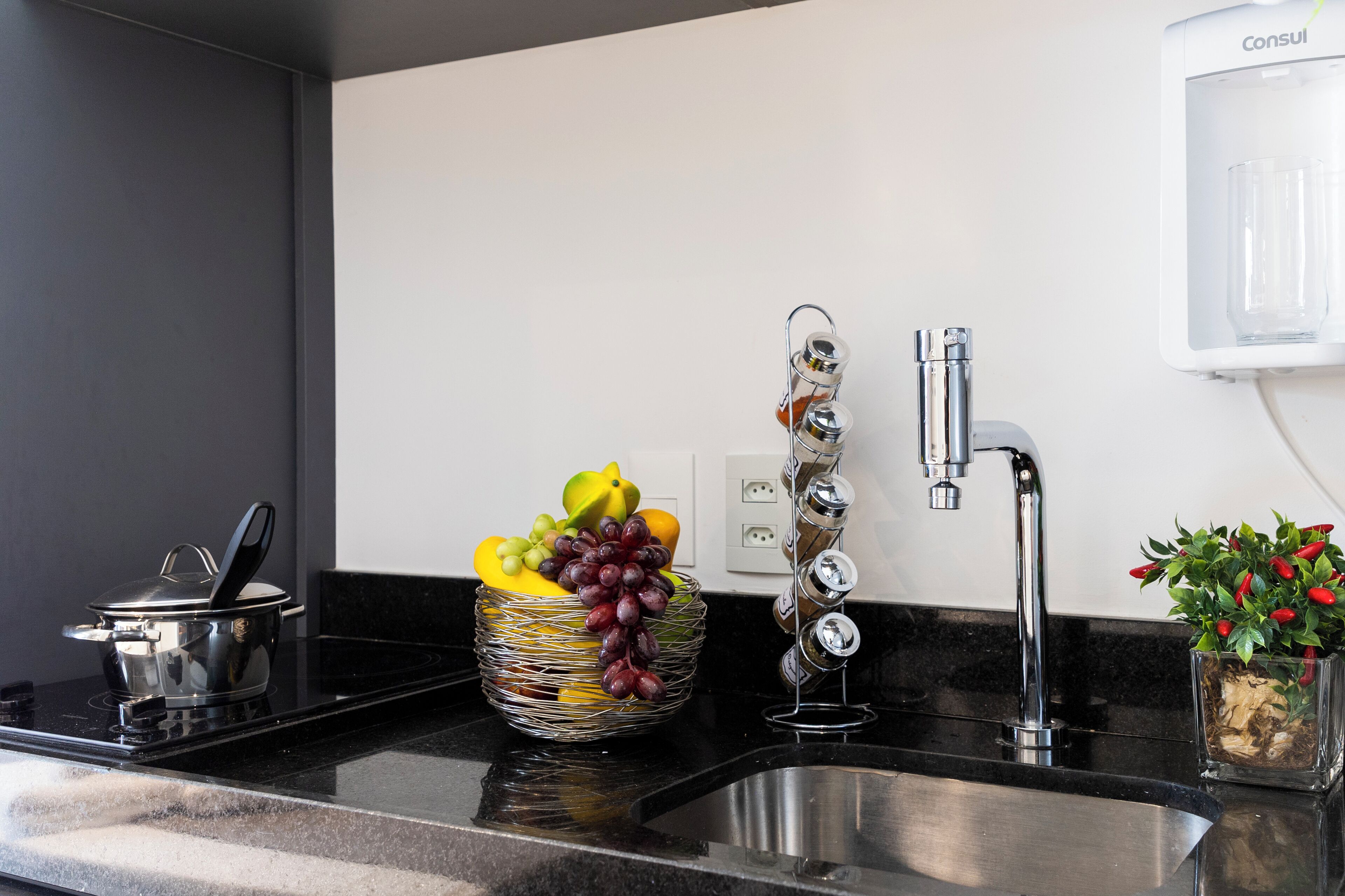 Una cocina con un lavamanos de acero inoxidable, una cesta de frutas, una olla en la estufa y una planta en maceta.