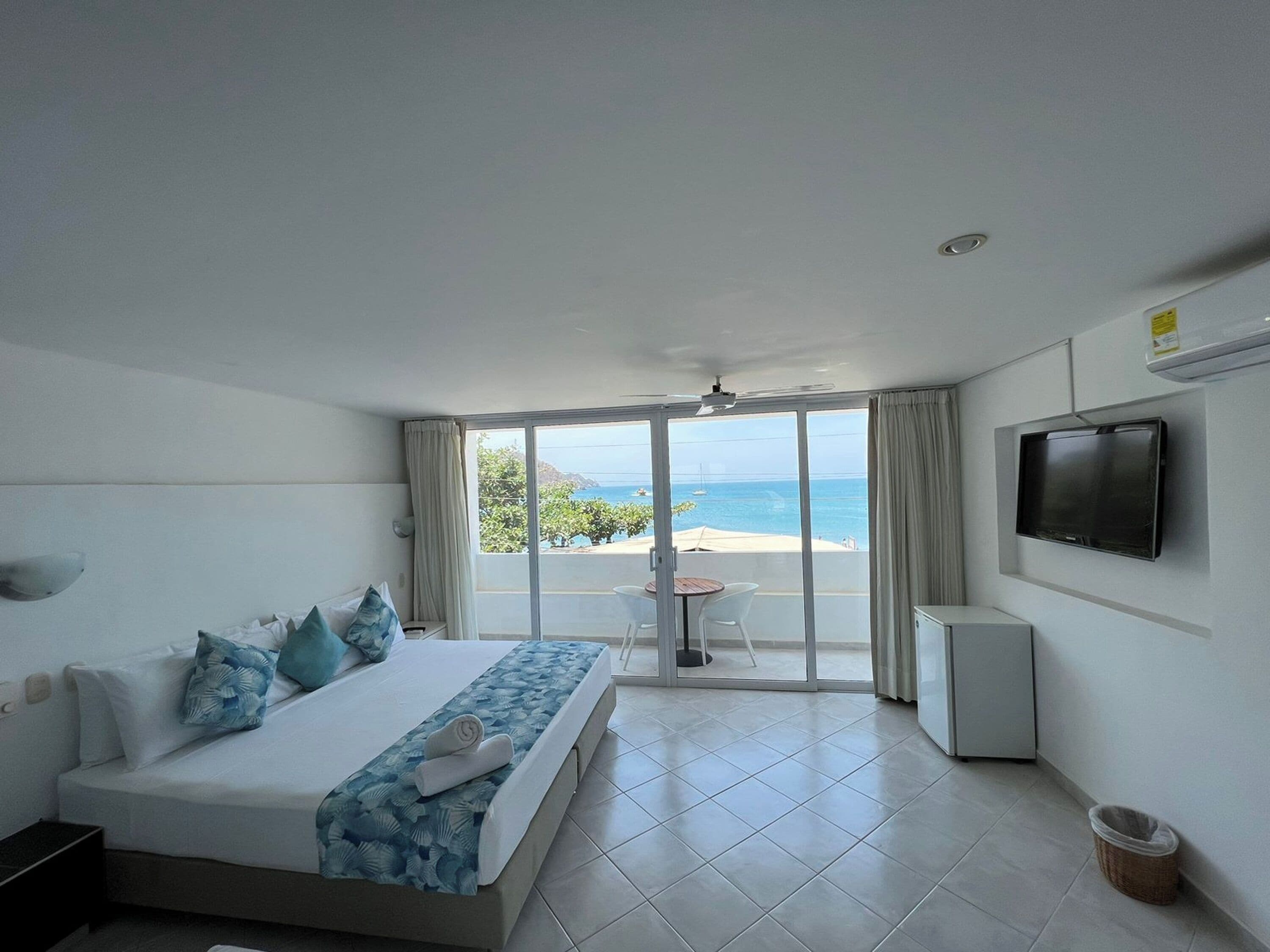A hotel room with a bed, a TV, a small table, and a view of the sea.