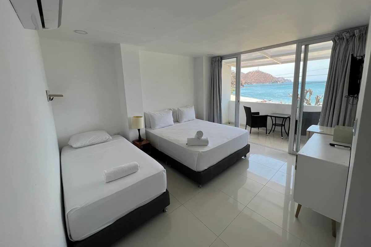 A hotel room with two beds, a desk, and a balcony with a view of the sea.
