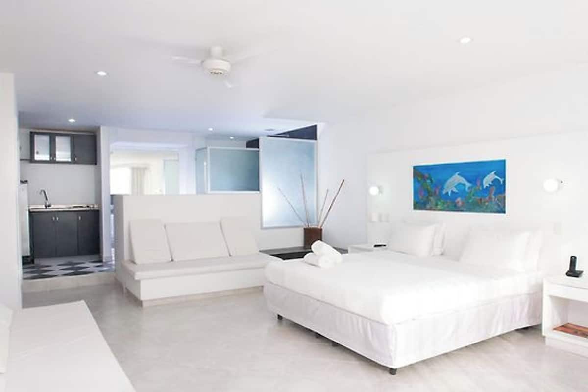 A modern hotel room with a large bed, a small seating area, a kitchenette, and a bathroom.