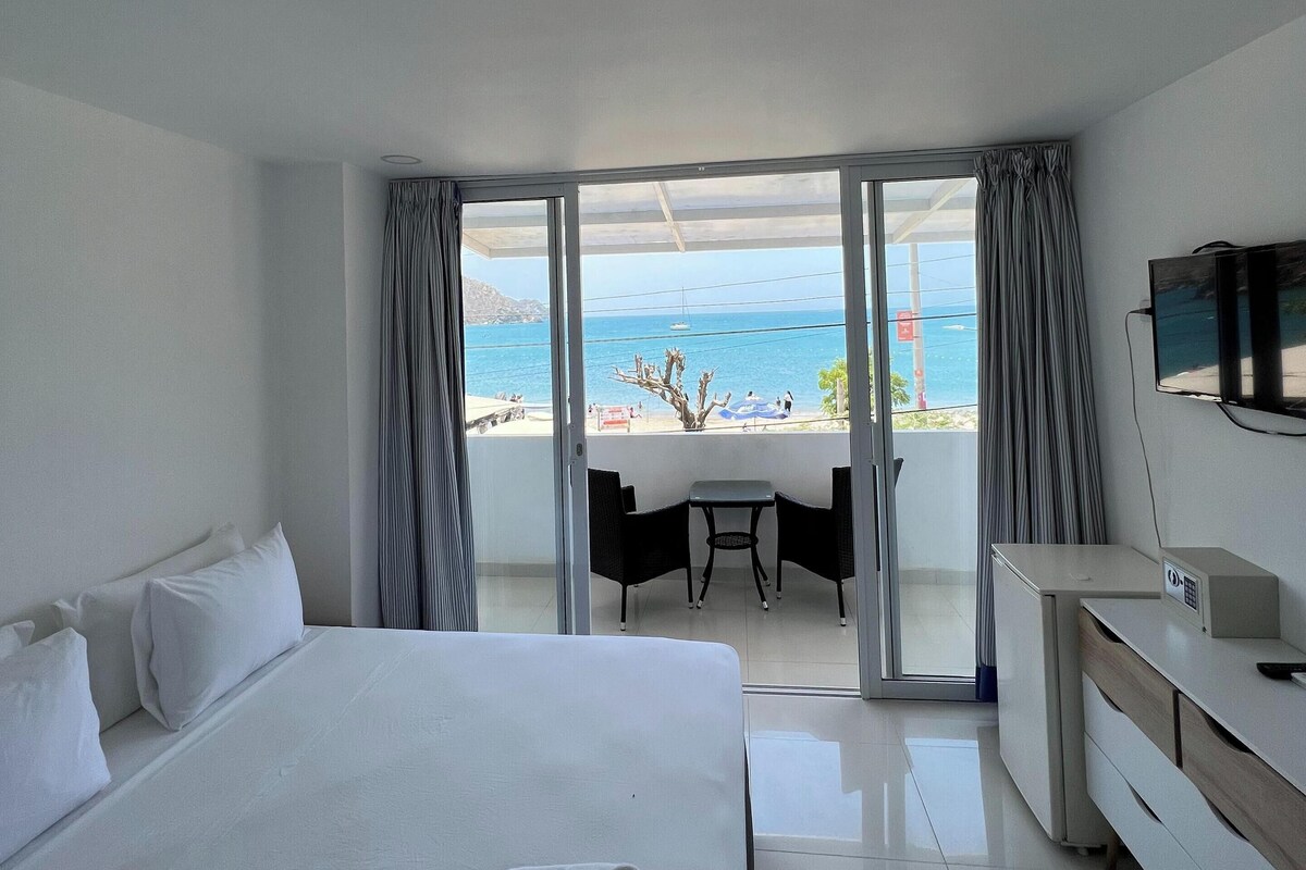 A hotel room with a bed, a desk, a chair, and a TV. There is a balcony with a view of the sea.