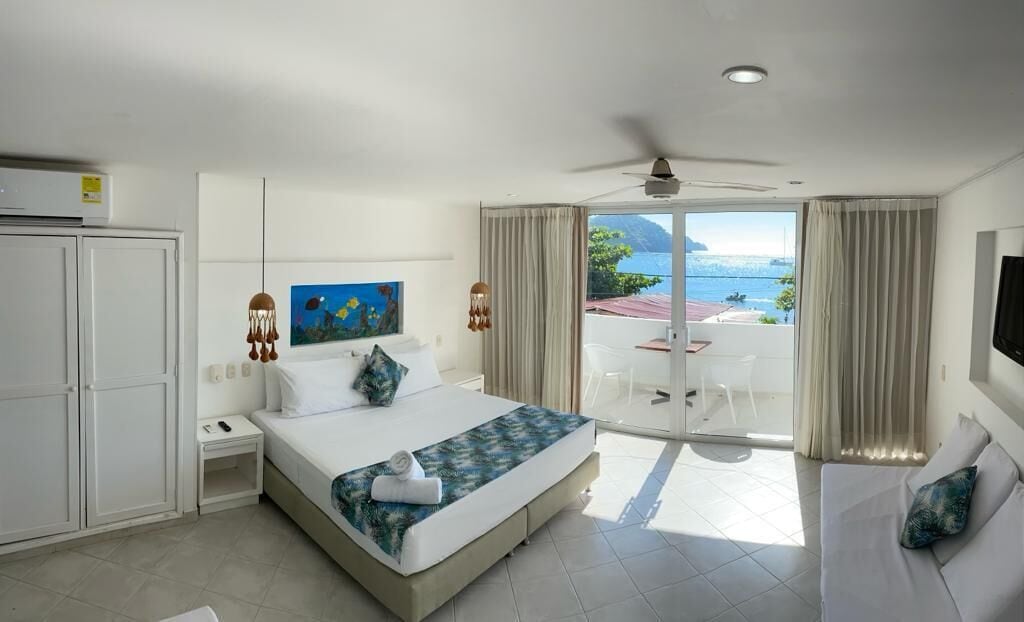A hotel room with a bed, a desk, a chair, a TV, and a balcony with a view of the sea.