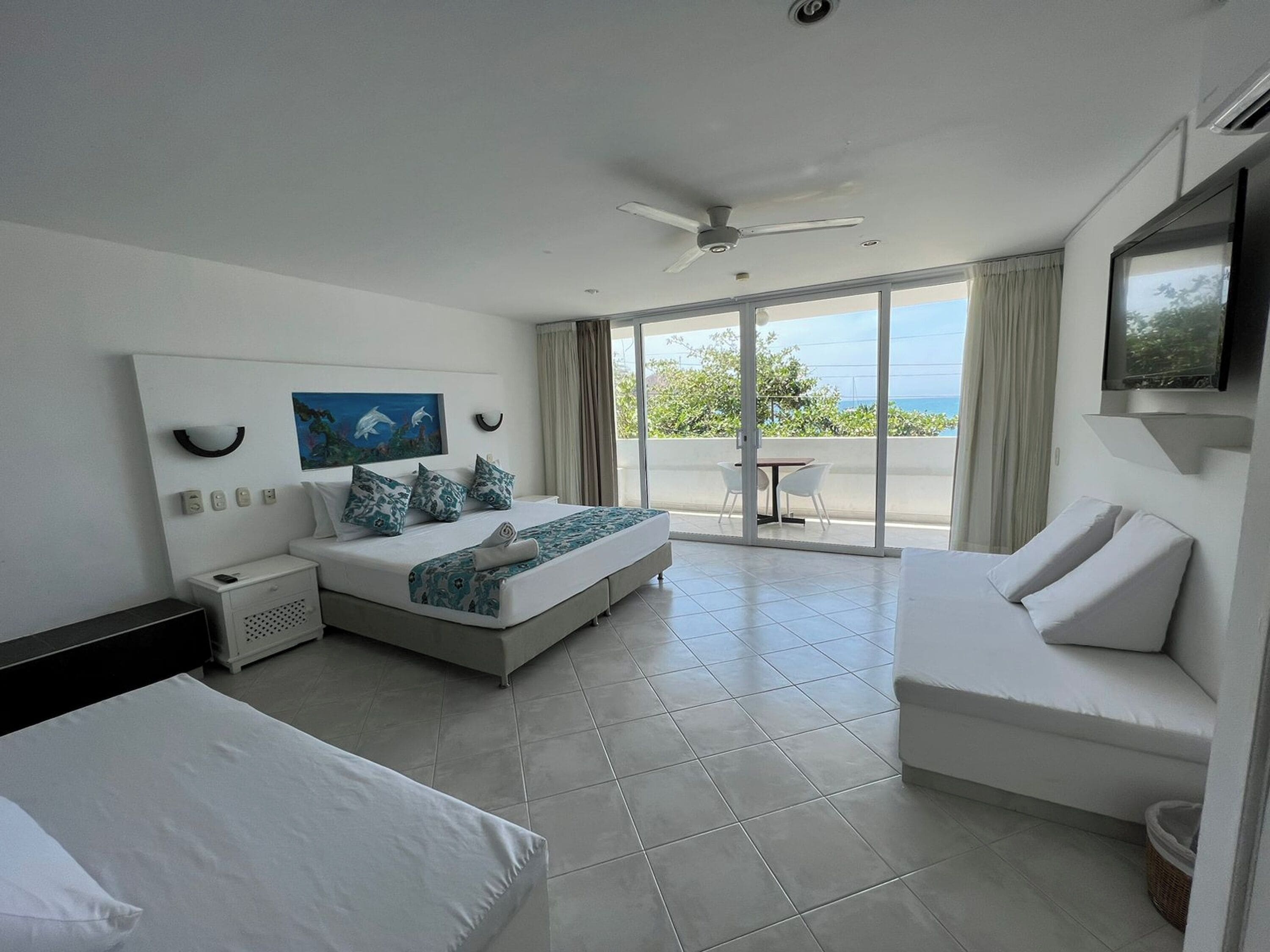 A modern hotel room with a large bed, a sofa, and a balcony with a view of the sea.