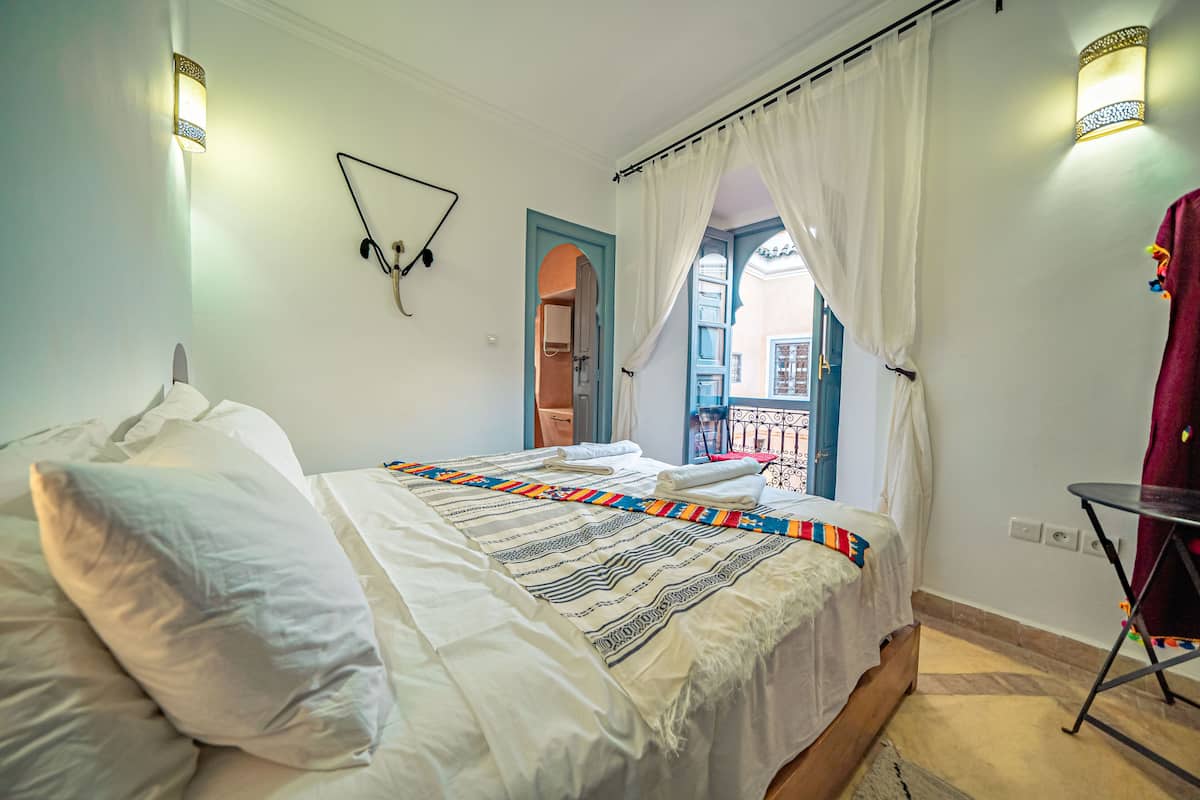 A bed with white linens, a colorful striped blanket, and a white pillow. There are two wall-mounted lamps, a black headboard, and a red towel on a chair.