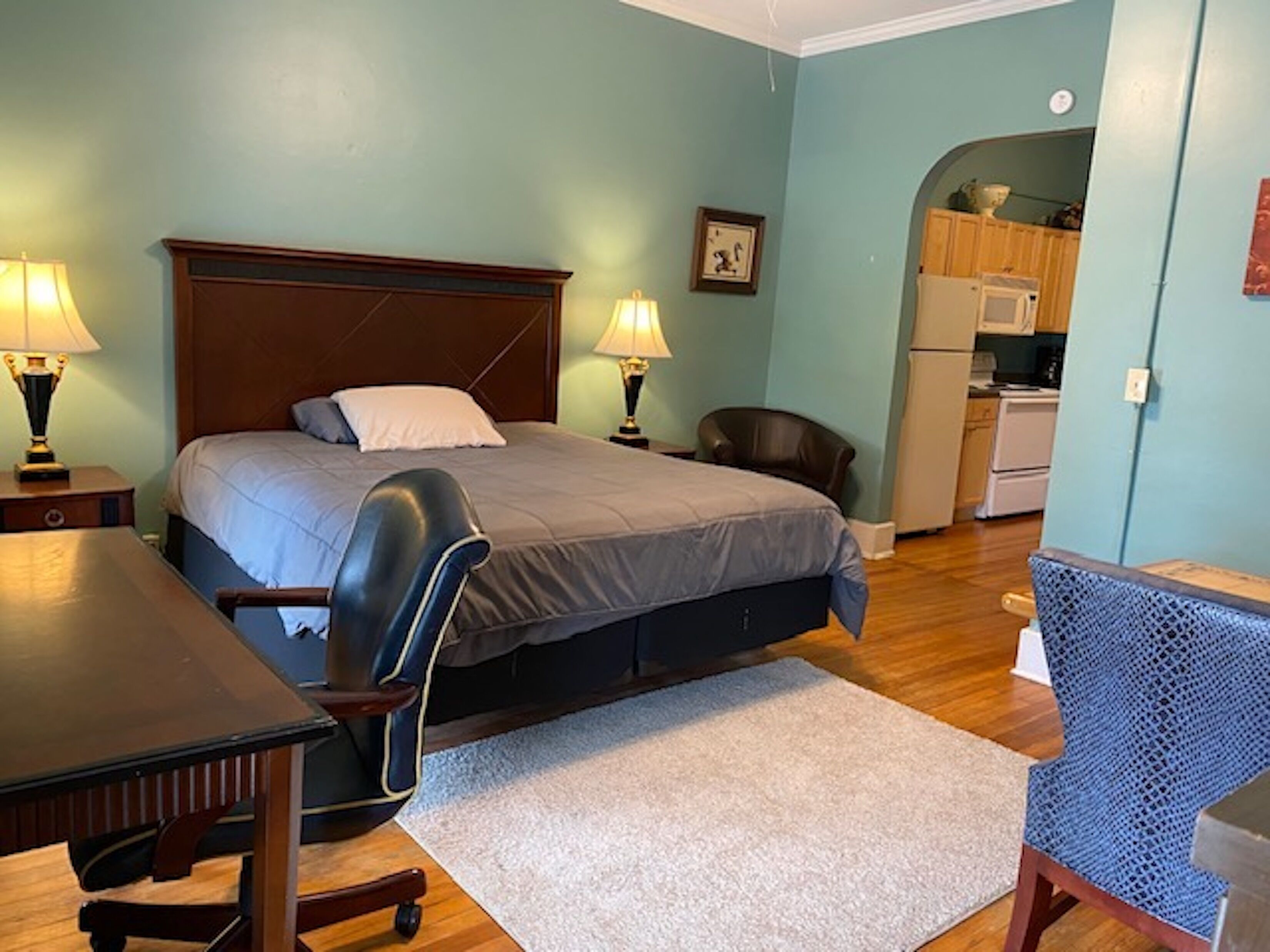 A bedroom with a bed, bedside tables, a desk, a chair, and a kitchenette in the background.
