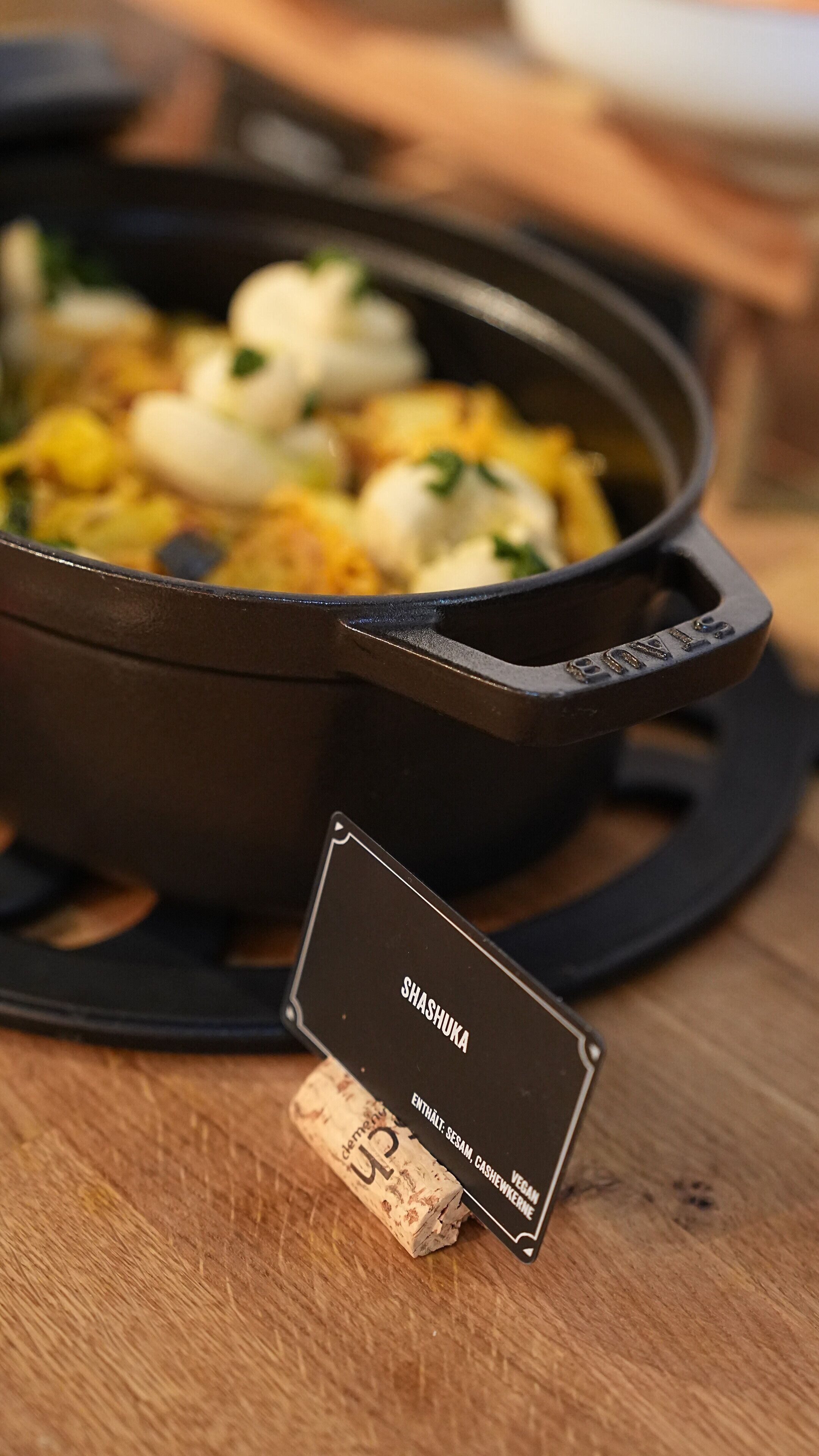 Eine schwarze gusseiserne Pfanne mit dem Aufdruck "SHASHUKA" und "ENTHALT: SESAM, CASHEWNERNE VEGAN".