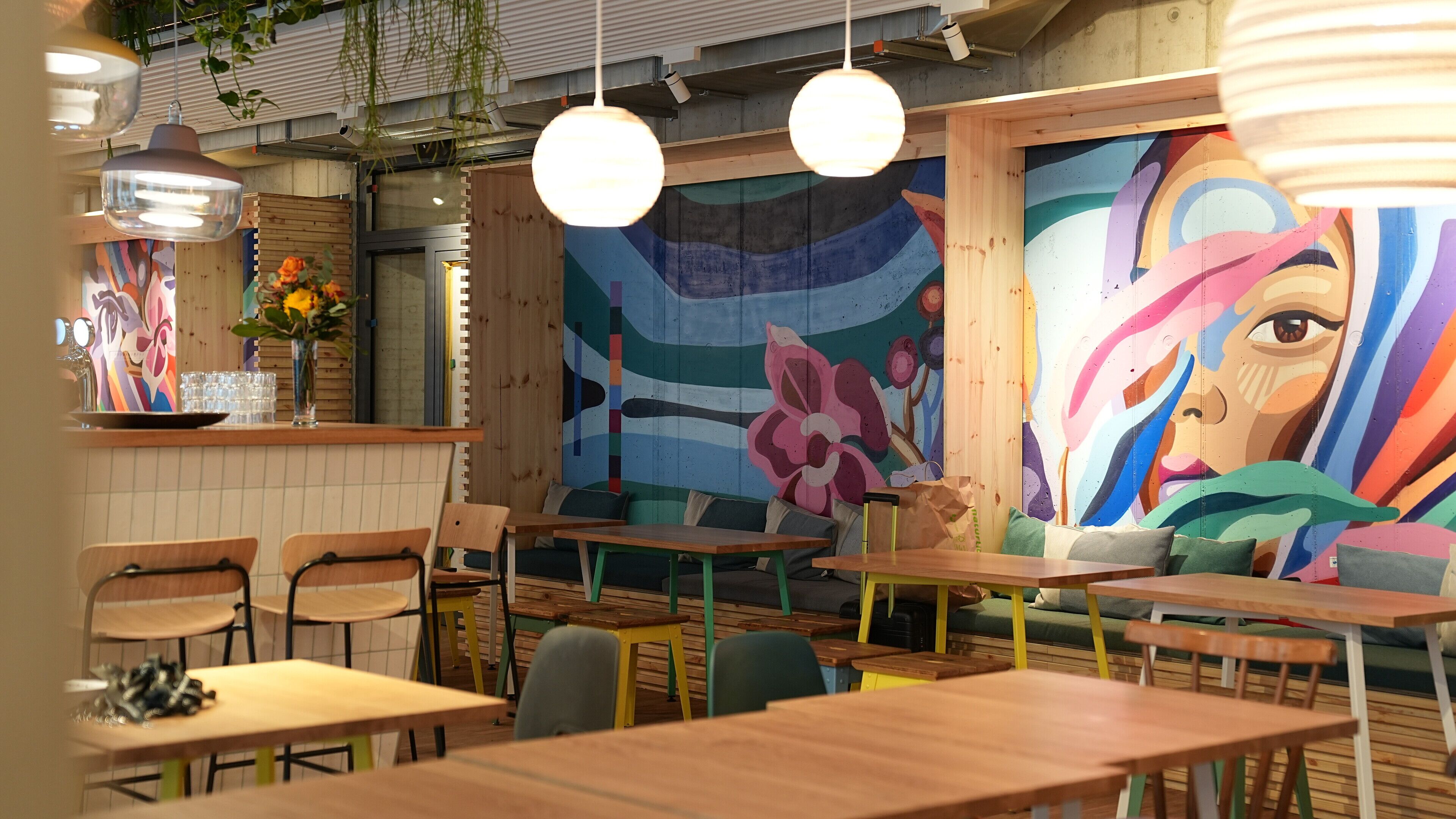 L’intérieur d’un restaurant est agrémenté de tables et de chaises en bois, d’une fresque murale, de luminaires suspendus et d’un comptoir orné d’un vase de fleurs.