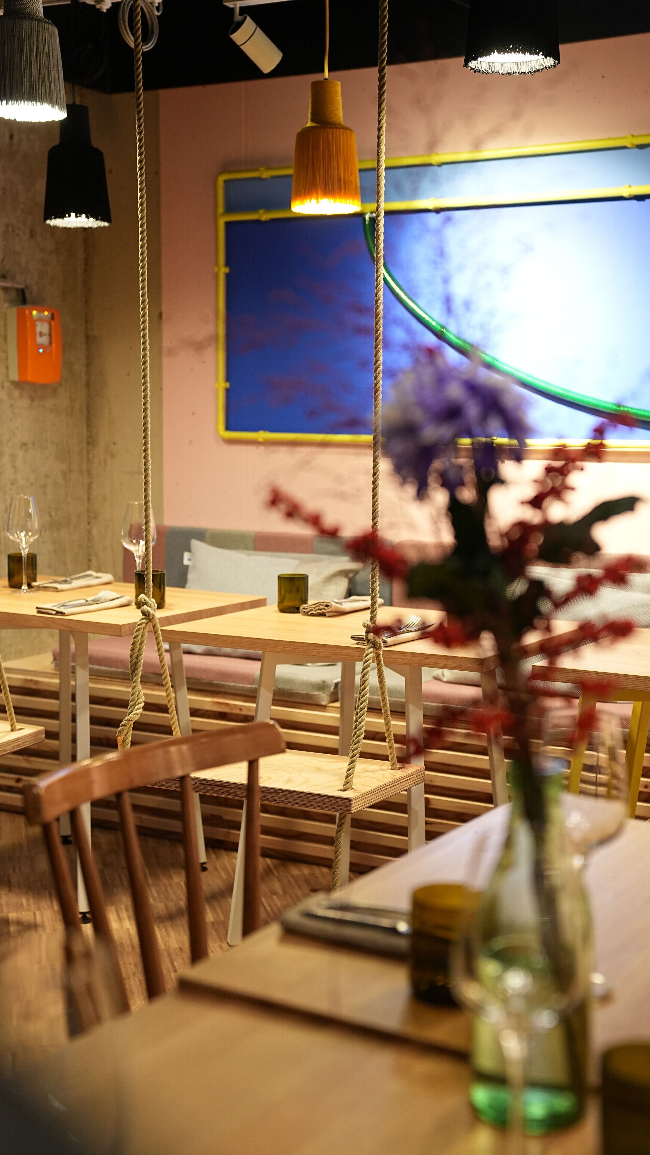 Interior de restaurante con mesas y sillas de madera, luces colgantes y un cuadro abstracto grande en la pared.