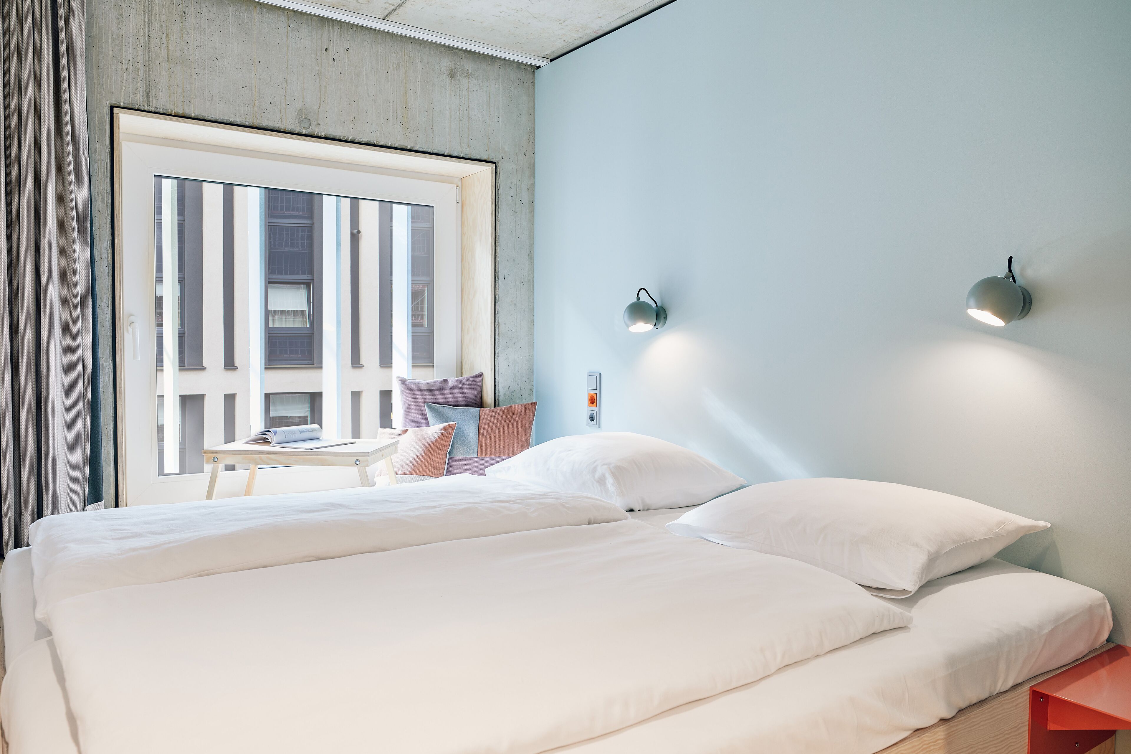 A hotel room with a large bed, a small table with a chair, and a window with curtains.