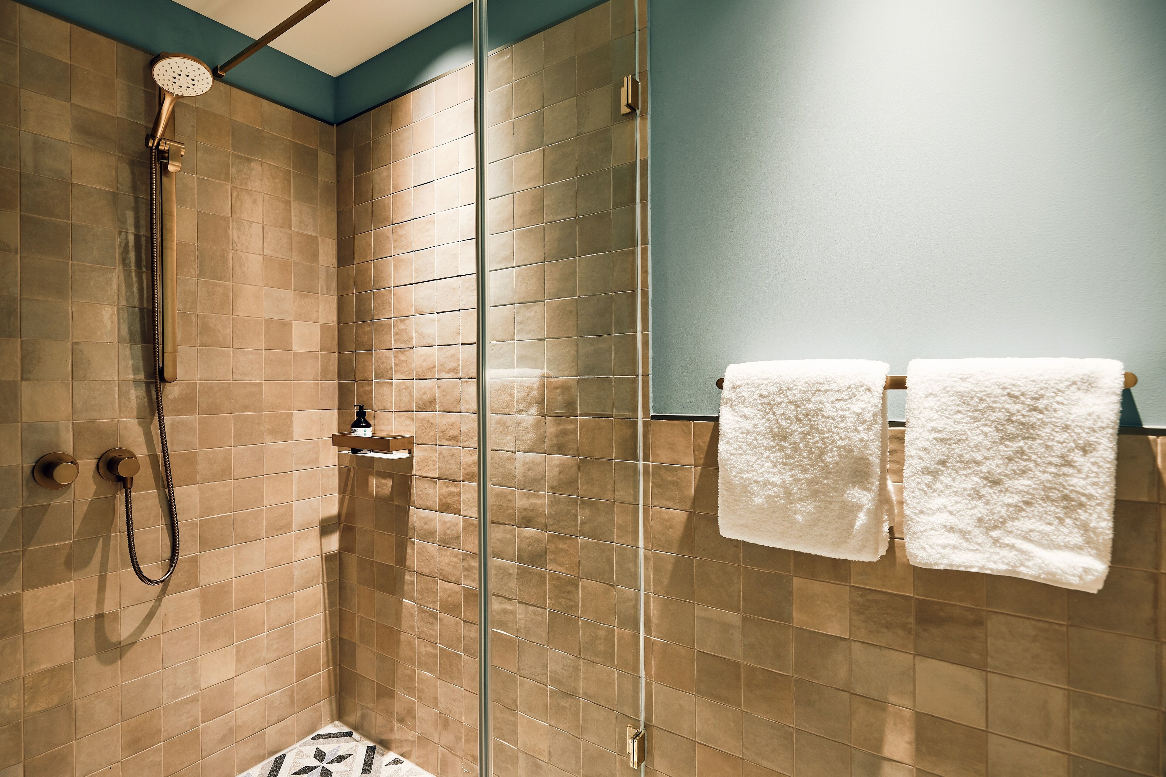 A glass-enclosed shower with beige tiles, a handheld showerhead, and two white towels on a rack.