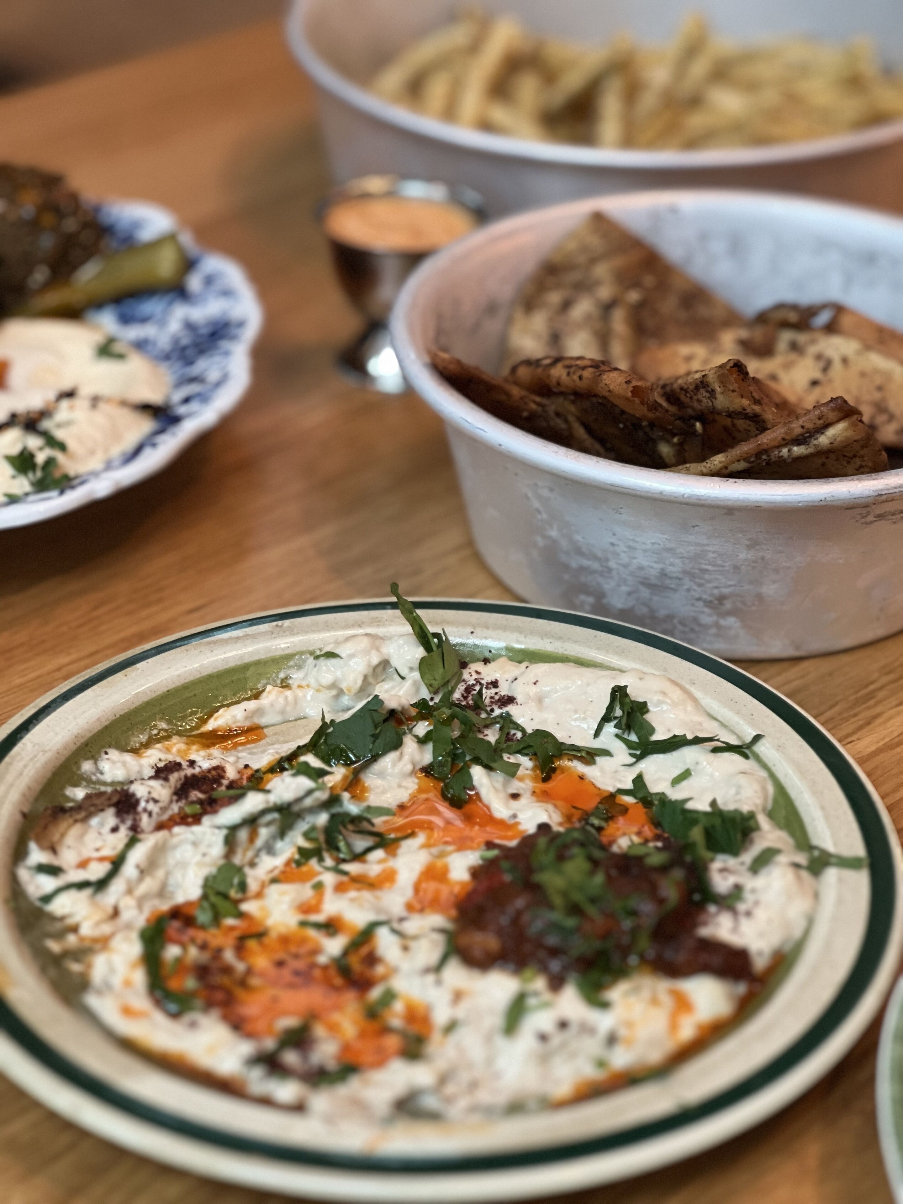 A table with various dishes including a bowl of hummus garnished with herbs and spices, a plate of fries, and a container of roasted potatoes.