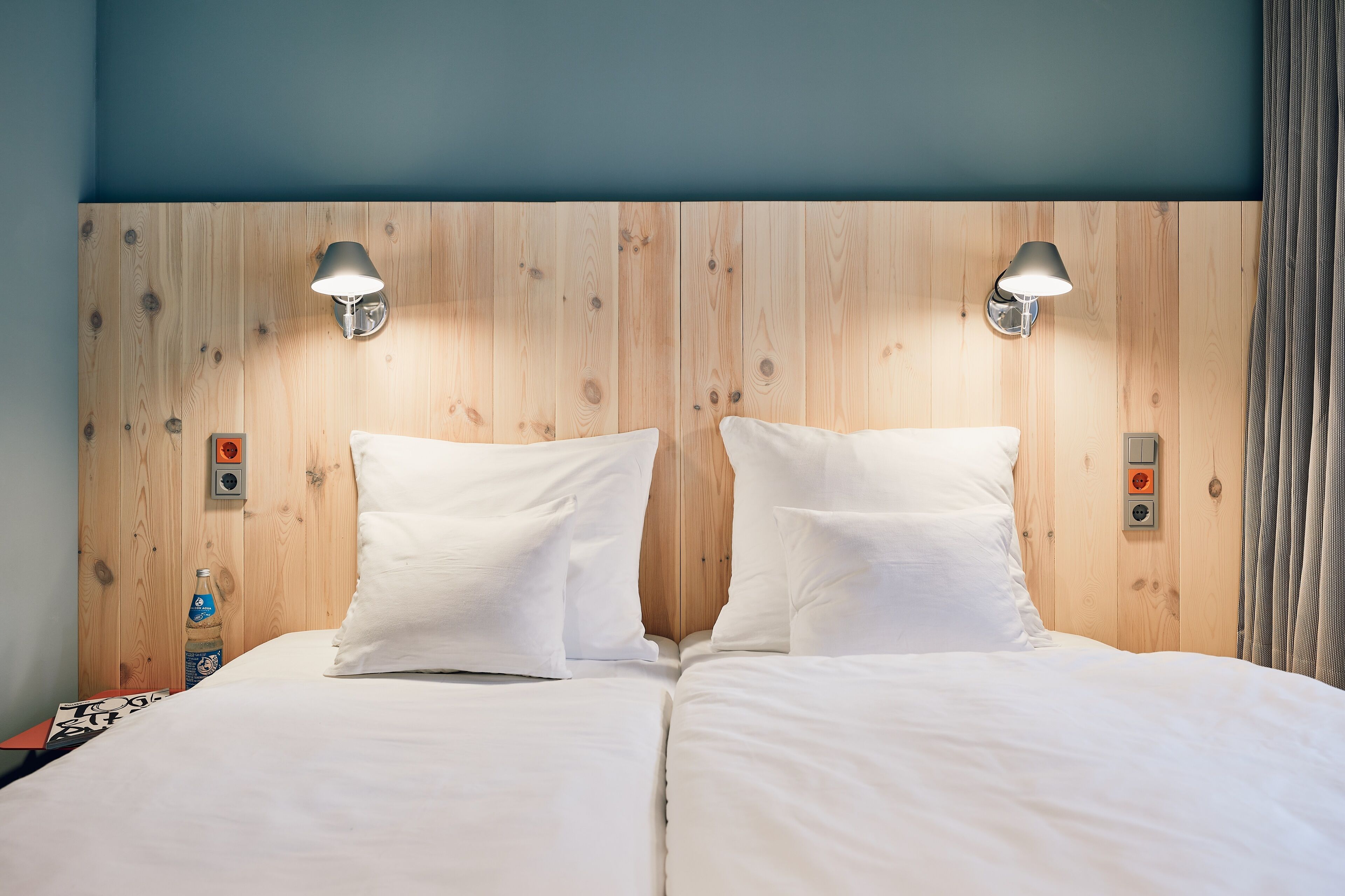 A double bed with white linens, two wall-mounted lamps, and a wooden headboard.