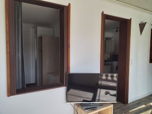 Ein Zimmer mit Fernseher auf einem Holztisch, einem Fenster mit Vorhängen und einer offenen Tür zu einer Küchenzeile.