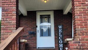 Una casa de ladrillo con una puerta blanca y un cartel que dice 'WELCOME'. Hay escalones que conducen a la puerta y una barandilla de madera a la izquierda.
