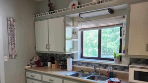 Una cocina compacta con armarios blancos, encimera negra, ventana con vistas a la vegetación y un felpudo negro en la entrada.