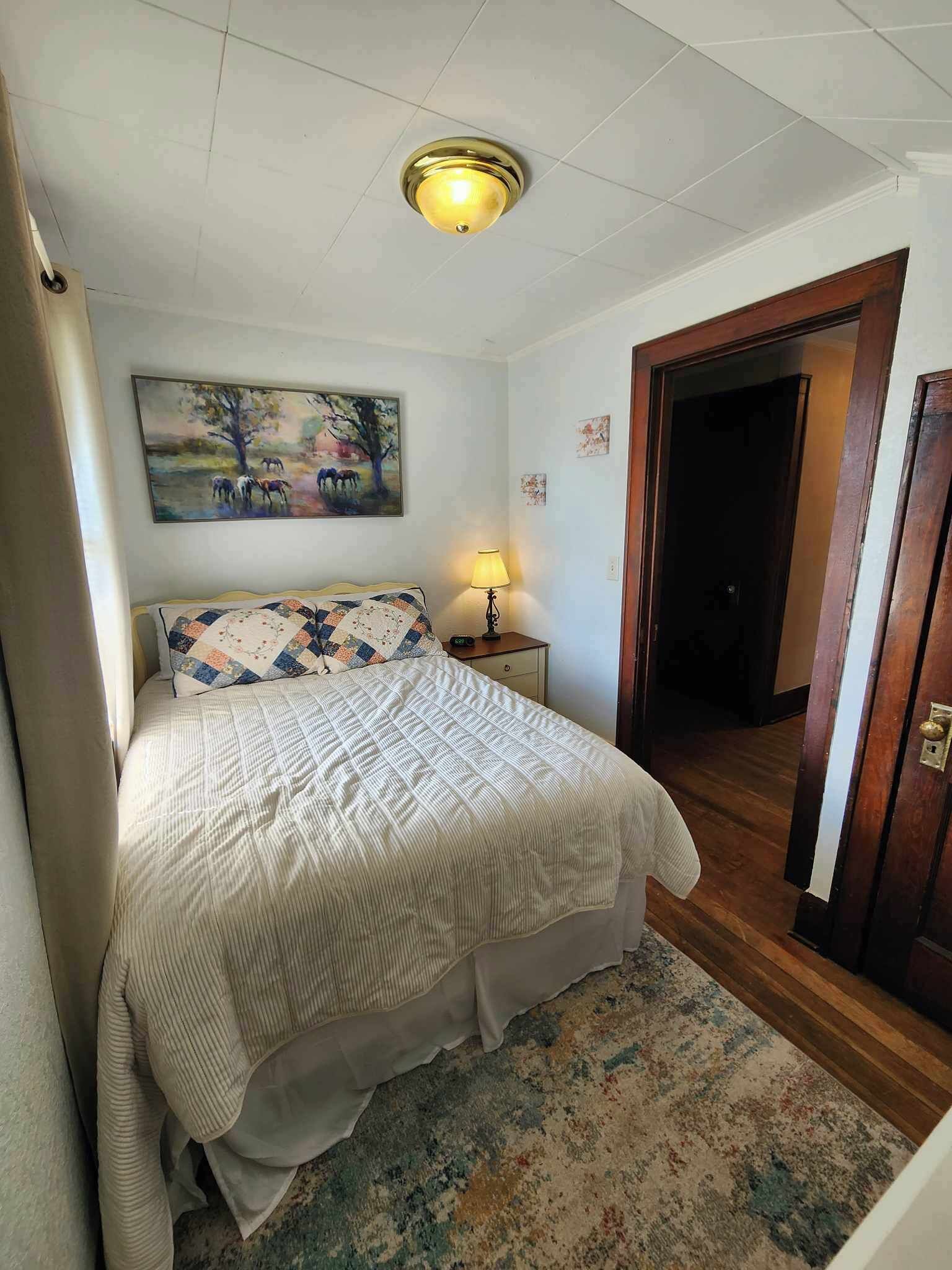 Ein Schlafzimmer mit Bett, Nachttisch, Lampe, einem Gemälde und einer Tür.