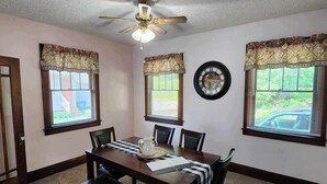 Un comedor con una mesa de madera, sillas, un banco, un ventilador de techo y tres ventanas adornadas con cortinas florales.