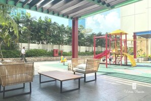 Relax in a serene outdoor space with a covered seating area and lush greenery, featuring a children's play structure and a large building in the background.