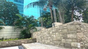 A stone wall with a metal railing, surrounded by greenery and palm trees, with a modern building in the background. - Impressive City View Suit for 4 Persons (West Jakarta City)