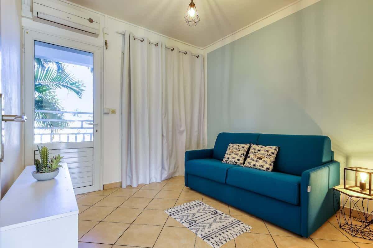 Ein Wohnzimmer mit einem blauen Sofa, einem weißen Beistelltisch mit Pflanze, einem gemusterten Teppich und einem Balkon mit Vorhängen.