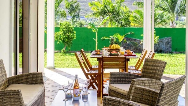 Une terrasse couverte, meublée de meubles en osier, avec une table à manger dressée avec des assiettes, des verres et une bouteille, et une vue sur un paysage verdoyant, avec des arbres et un mur de pierre.