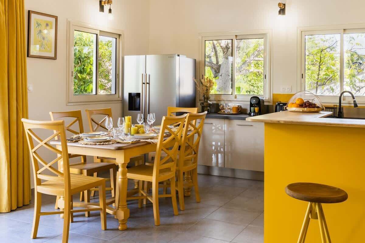 Une cuisine lumineuse aux touches de jaune, un coin repas avec des chaises en bois et de grandes fenêtres offrant une vue sur la verdure.