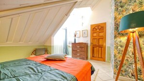 Ein Schlafzimmer mit schrägen Wänden, einem Bett mit einer grünen und orangefarbenen Bettdecke, einer Holzkommode, einer Tür und einer Wand mit tropischem Tapetenmuster.