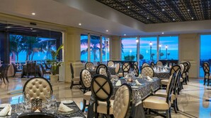 Un comedor amplio con varias mesas y sillas, ventanas grandes con vistas a palmeras y al océano, y un techo decorativo.