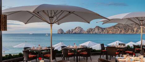 Un área de comedor al aire libre con mesas y sillas, sombrillas grandes y vistas al mar y acantilados rocosos.